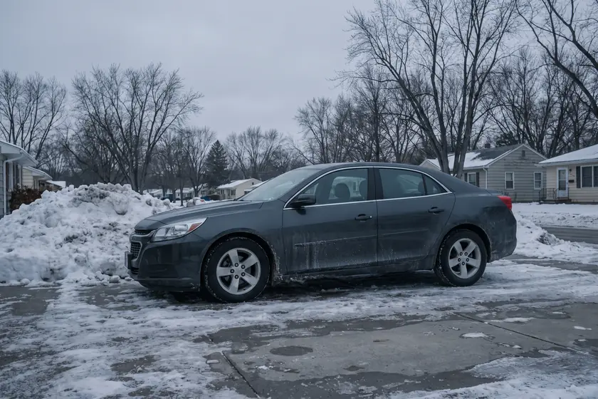 A grey sedan on a wet driveway beside a plowed snowbank in a Midwestern suburb
