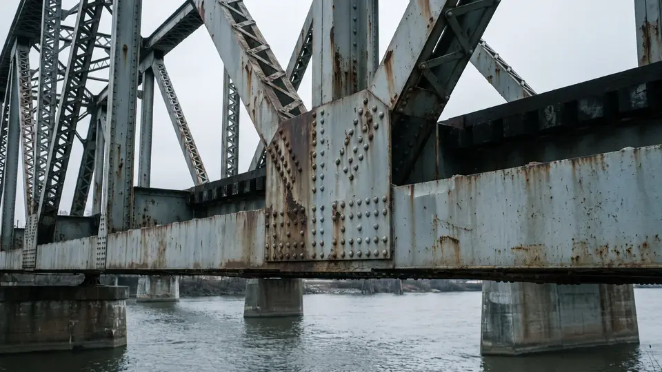 A riveted steel truss bridge over an industrial river under an overcast sky